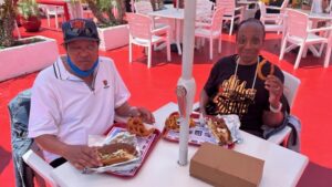 The picture shows two people sitting at a table in what appears to be an outdoor dining area of a restaurant. The area has a vibrant atmosphere with a bright red floor and white furniture. There are also some plants and flowers in the background. On the left, there is a man wearing a white polo shirt, a cap with a Chicago Bears logo, and sunglasses. He has a blue face mask pulled down to his chin and is sitting in front of a tray with a sandwich and onion rings. On the right, there is a woman wearing a black t-shirt with gold lettering, and she is holding up an onion ring with a smile on her face. She also has a tray in front of her with a sandwich and fries. In the middle of the table, there is a tall white umbrella stand. The table also has some trays with food and a brown box. The overall mood of the picture seems cheerful and relaxed, as if they are enjoying a nice meal outside.