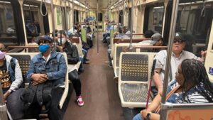 The picture shows the interior of a public transport train, possibly a subway or metro. The train car is well-lit and has a long aisle with rows of seats on both sides. The seats are patterned with dots and are in shades of brown and beige. There are metal poles and handles for standing passengers to hold onto. There are several passengers in the train car, most of whom are wearing masks, which suggests that the picture was taken during the COVID-19 pandemic. The passengers are of various ages and ethnicities. Some are sitting while others are standing. They are dressed in casual attire, including jeans, t-shirts, and hats. In the foreground, there is a man wearing sunglasses, a blue mask, and a denim jacket. He is sitting and holding a backpack on his lap. Next to him, there is a woman wearing a purple mask. On the right side of the picture, there is a woman with braided hair wearing glasses and a striped shirt. She is sitting and holding onto a pole. Behind her, there is a man wearing a hat with the letters "LA" on it, sunglasses, and a checkered shirt. The atmosphere in the train car seems calm and quiet, with passengers minding their own business.