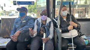 The picture shows three people sitting inside a bus. They are seated in an area that is reserved for mobility devices, as indicated by a sign above them. From left to right: 1. The first person is a man wearing a denim jacket, a cap, and a blue face mask. He is looking to his left and has a black bag on his lap. 2. The second person is a woman wearing sunglasses, a purple face mask, and a denim jacket over a black and white outfit. She is holding a white cane with a curved handle and has a black bag on her lap. She is also wearing a necklace with white beads. 3. The third person is a woman wearing a black jacket, black pants, and a white face mask. She is holding a white cane with a straight handle and has a black bag with some items in it hanging on the armrest. The bus has large windows, and you can see the street, buildings, and trees outside. The seats in the bus have a pattern with circles and dots.